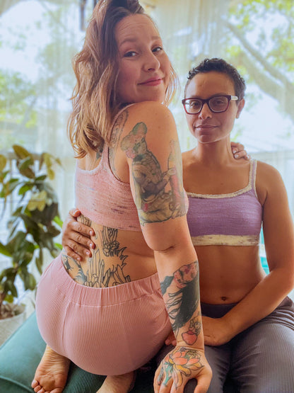 Sitting facing each other, Candace and Bess wears hand knit bralettes. Candace's is knit with light purple yarn and a contrasting white speckled band and straps. Bess's is knit with light pink speckled yarn, and she wears matching leggings. They both are turned to smile at the camera.