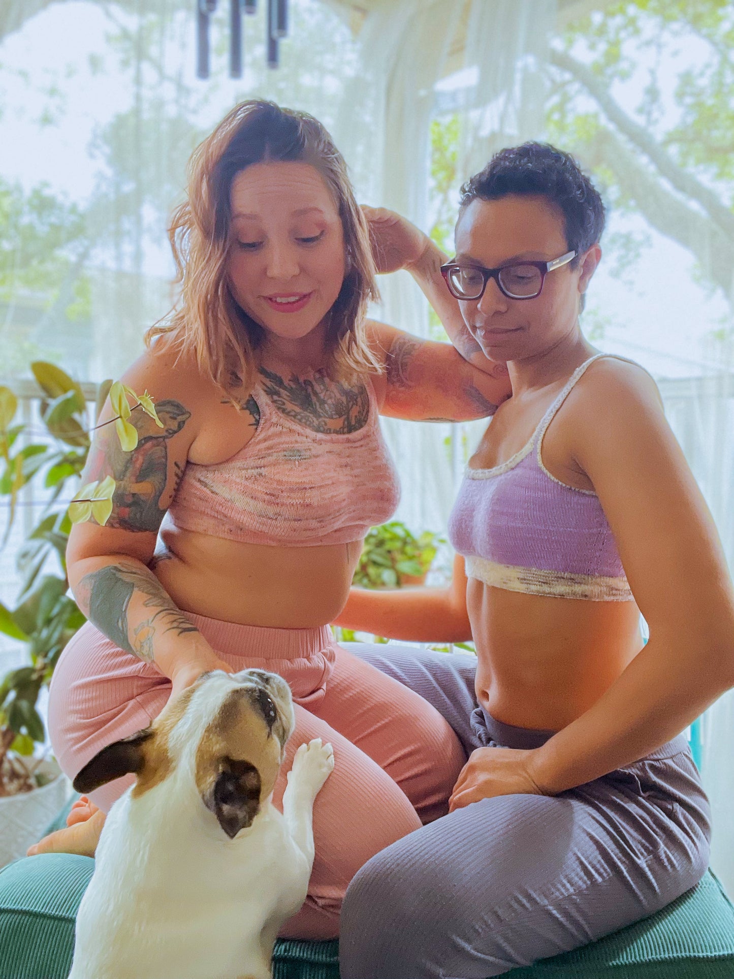 Bess and Candace kneel on a puff stool, looking down at a white and brown Frenchie. Bess and Candace each wear a well-fitted, hand knit bralette. Bess' is made with light pink speckled yarn, and Candace is made with light purple yarn and a white speckled band.