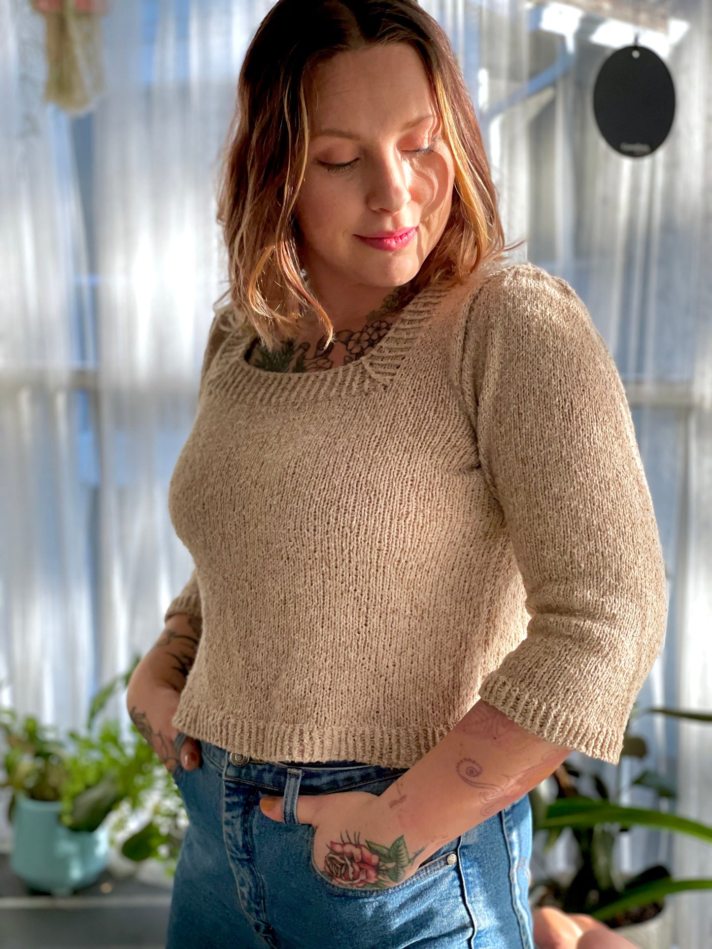 Bess stands, looking off camera. Her light beige sweater is knit with a square neck and 3/4 length sleeves, which she's paired with blue jeans.