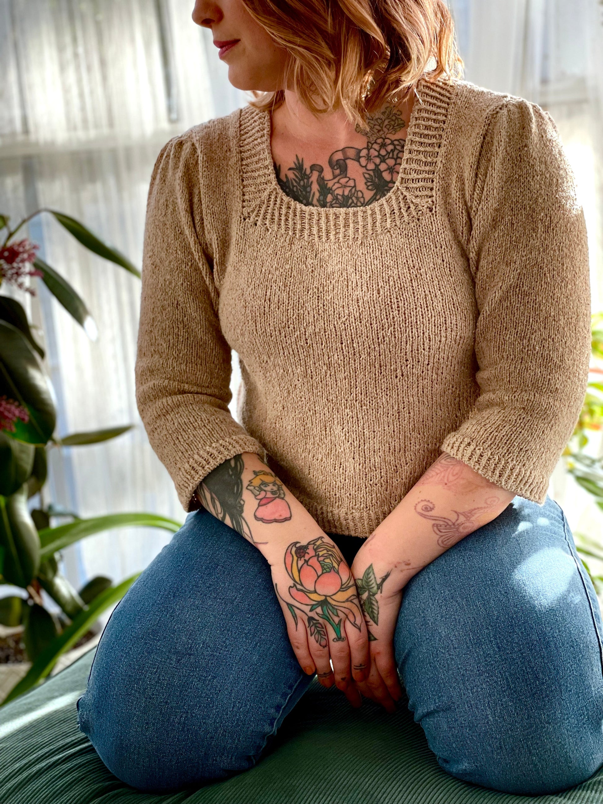 Bess kneels on a footstool, wearing a light beige sweater and blue jeans. The sweater is knit with a square neckline and 3/4 sleeves, with have been gathered at the top to create pleats.