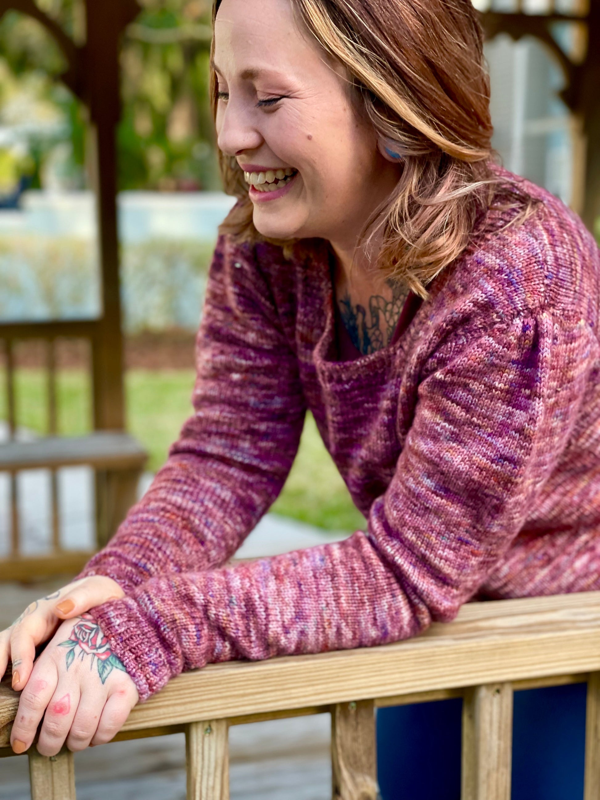 Bess laughs, leaning against a wooden deck railing. She wears a hand knit sweater, made from pink variegated yarn. The sleeves of the sweater have been gathered at the top to create pleats.
