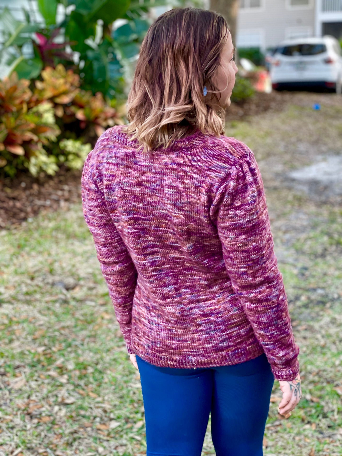 Seen from behind, Bess stands in a backyard. She wears a pink variegated sweater, knit with gathered and pleated sleeves, with a pair of blue leggings.