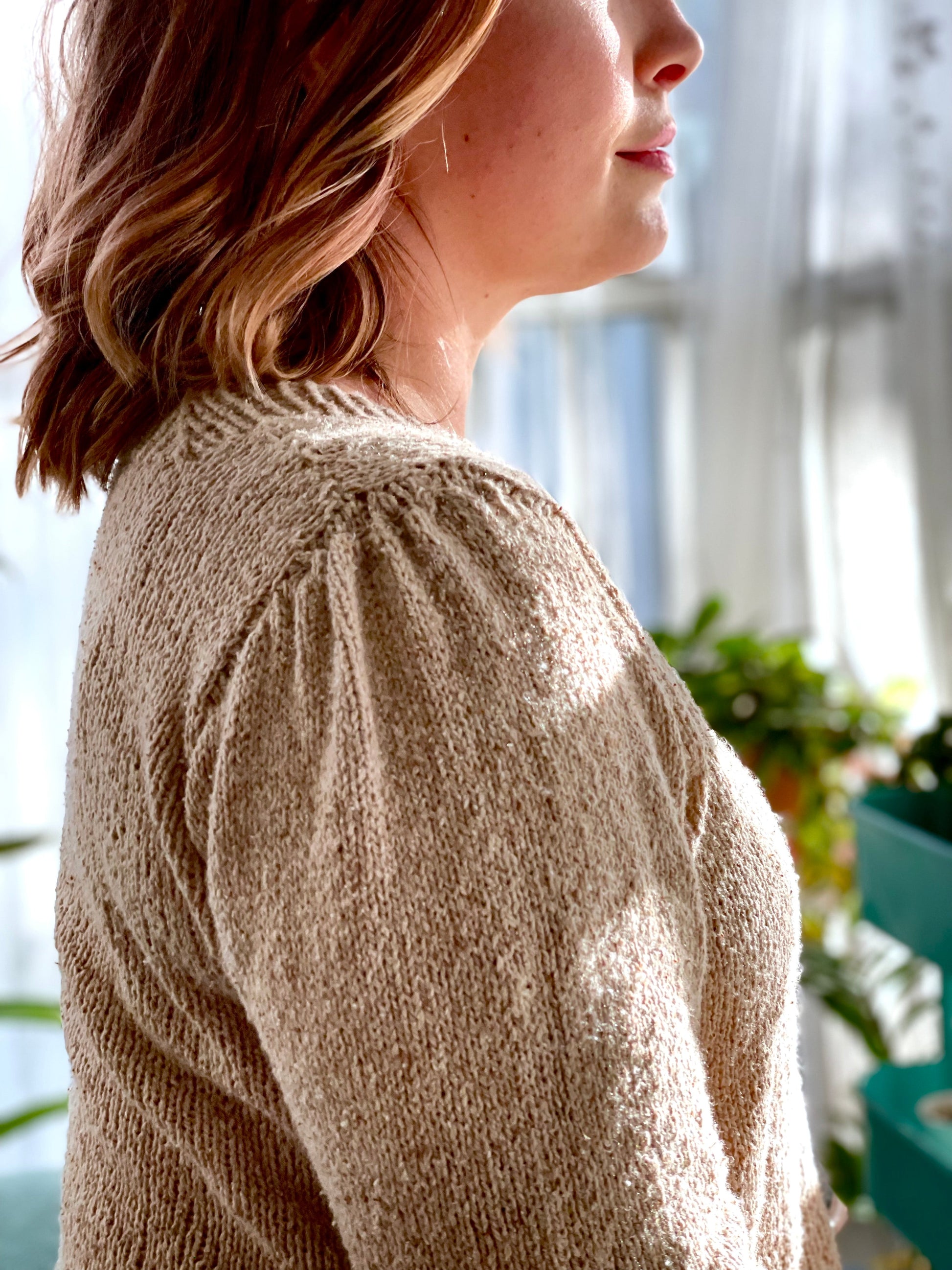 Bess, seen from the side, wears a light beige knit sweater - the Hypatia pullover. The sleeves feature a gathered seamed, creating pleats at the shoulder and upper arm.