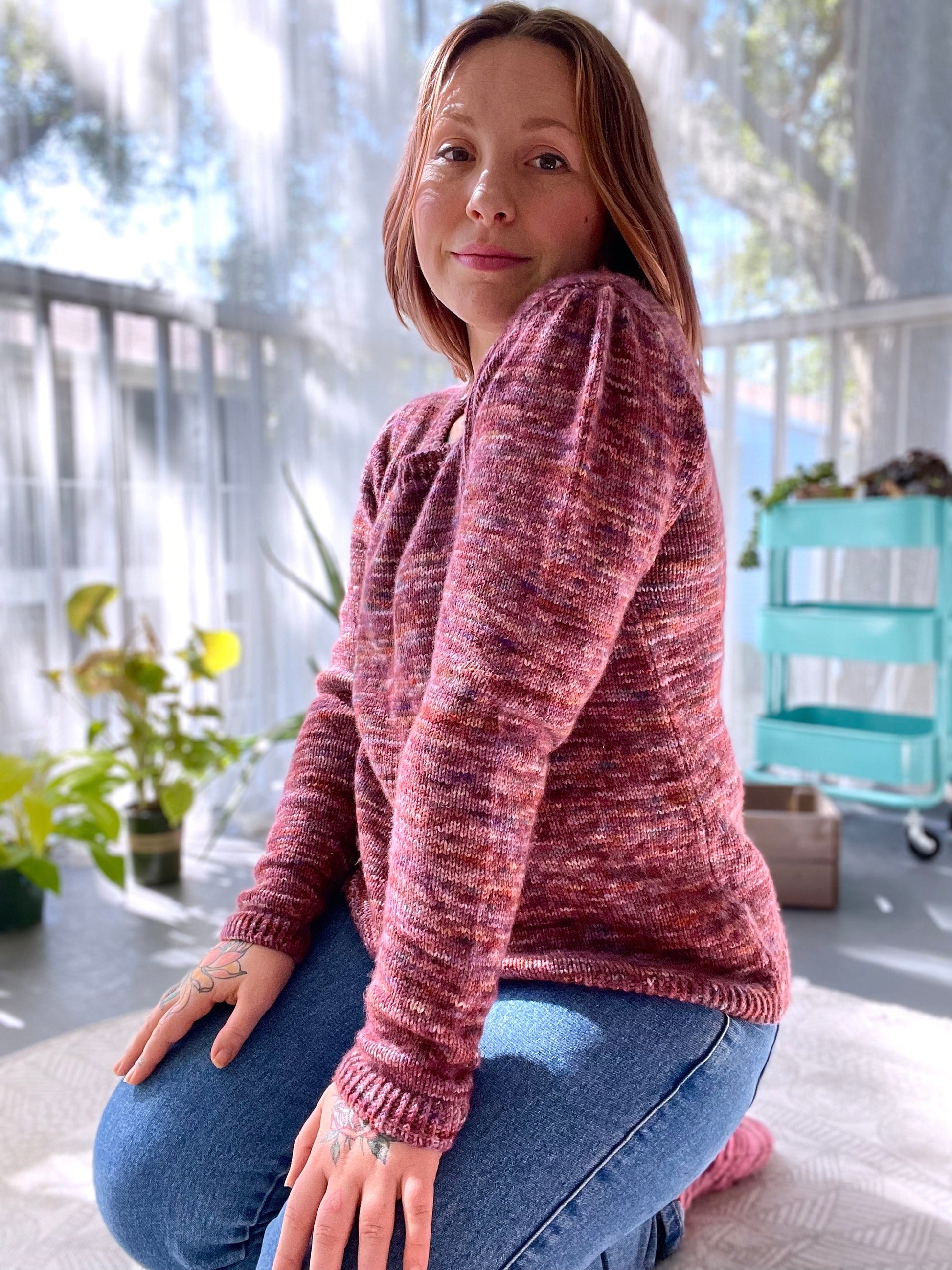 Seen from an angle, Bess kneels on the floor, smiling at the camera. She wears a pink variegated sweater with pleated ruffle sleeves.