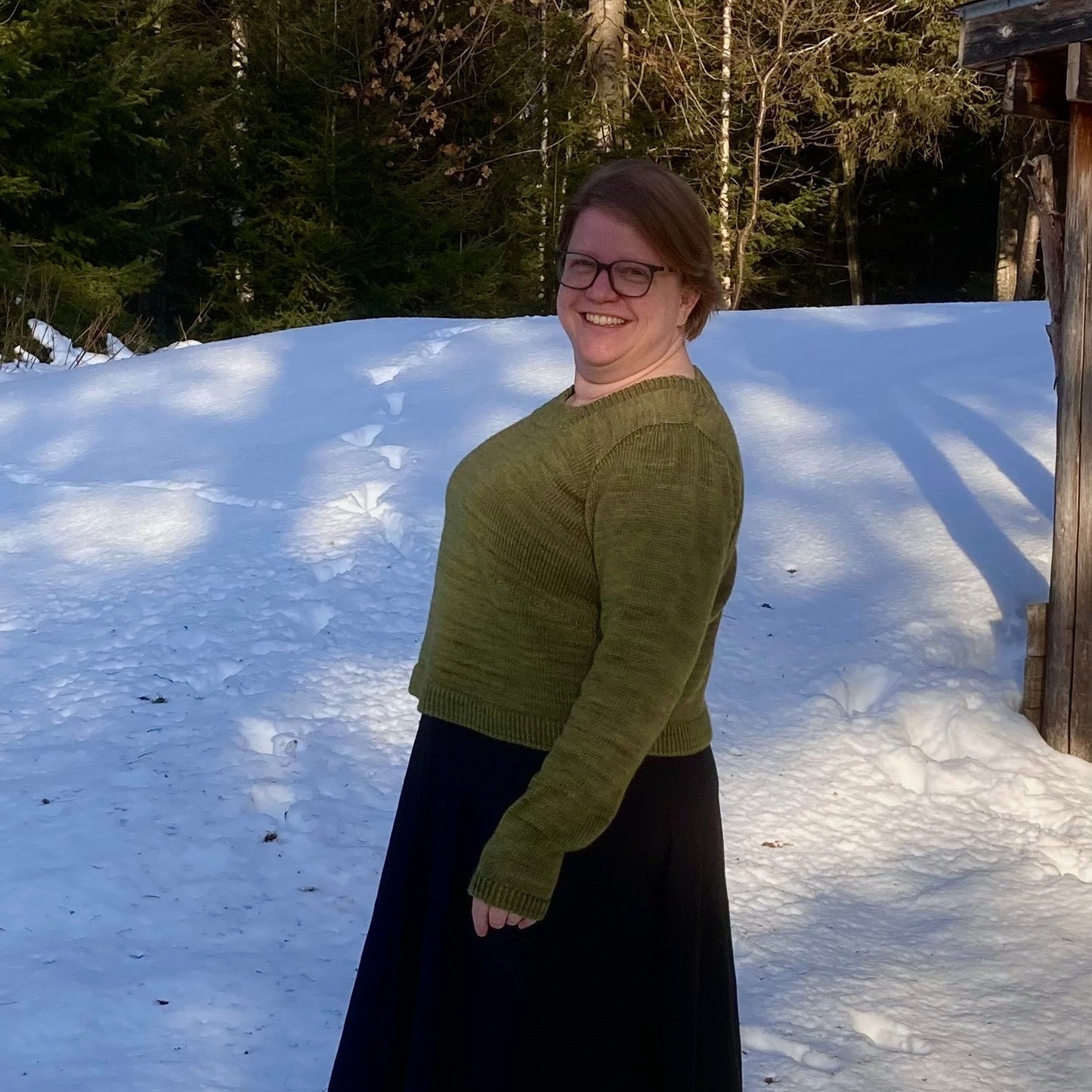 Seen from the side, a person wears a hand knit, green sweater, design with gathered sleeves, with a black skirt. They smile at the camera.