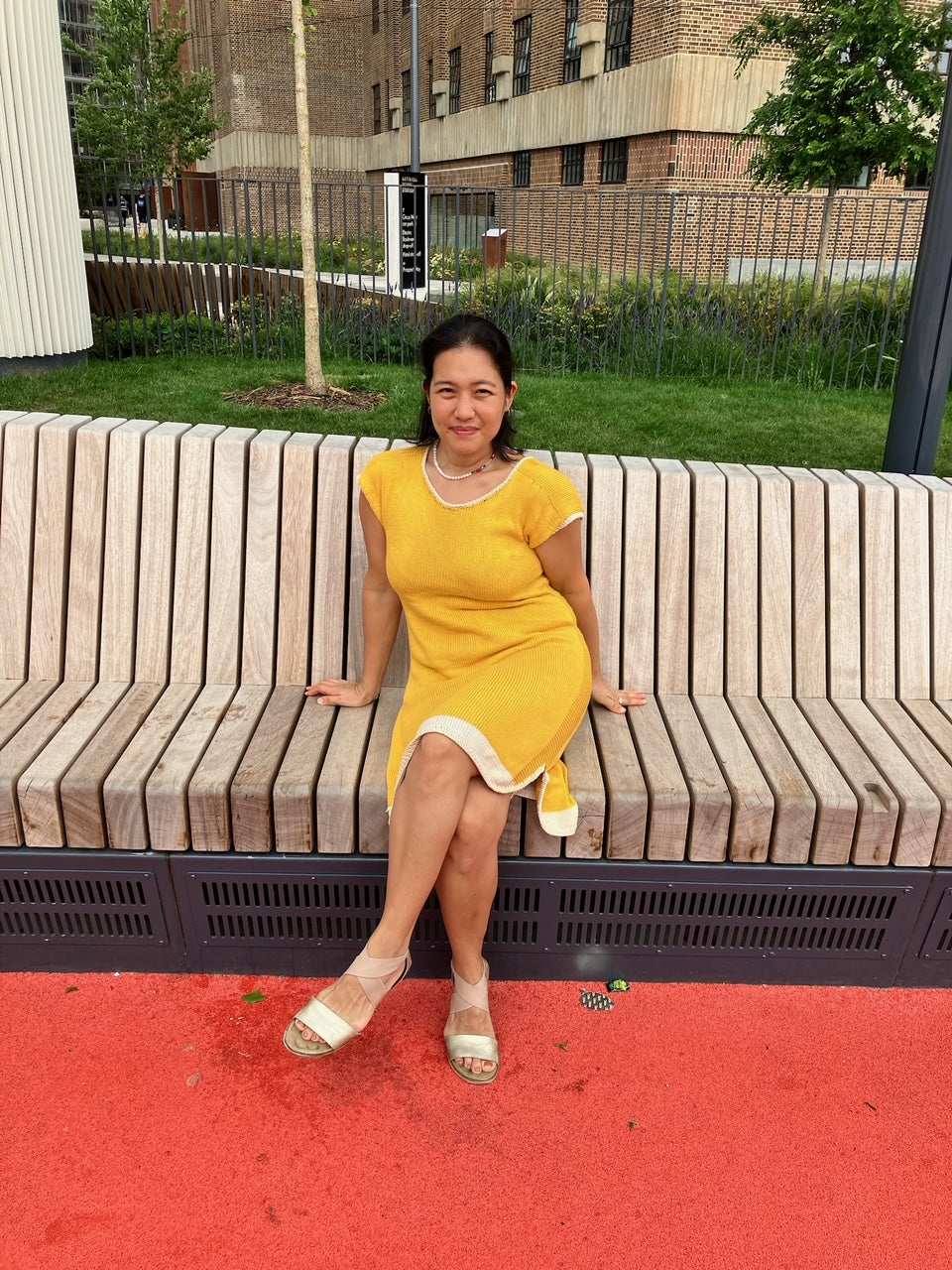 A person sits on a wooden bench, wearing a bright yellow and white hand knit dress with short sleeves.