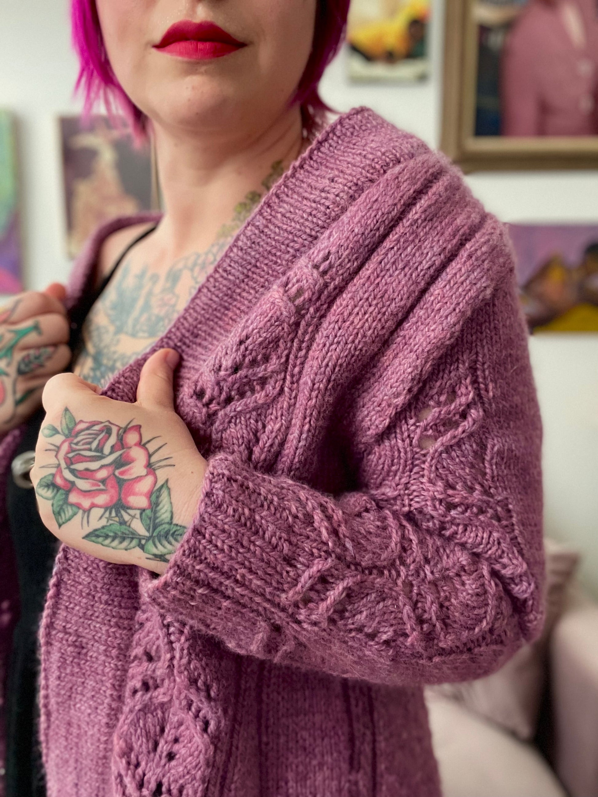 Seen from the neck down, Bess holds the ribbed edges of her light pink cardigan. Knit with bulky yarn, the cardigan features lace panels down the fronts and sleeves. Paintings hanging on a wall can be seen in the background.