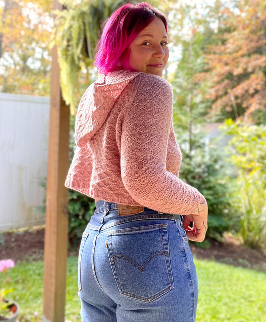 Bess, seen from behind, wears a light pink cropped hoodie with an all-over diamond pattern, paired with blue jeans. A backyard garden can be seen in the background.