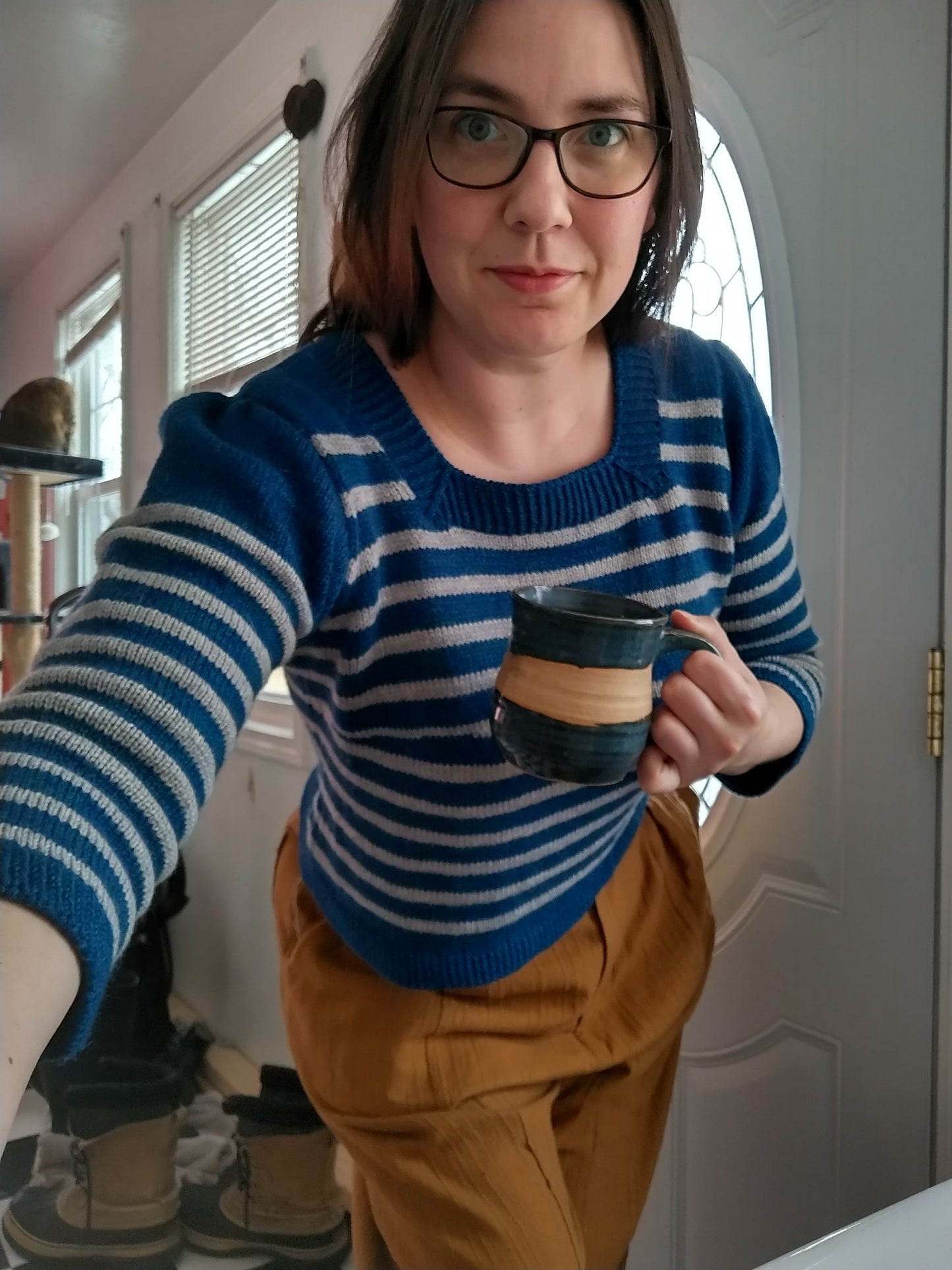 A person holds a mug, leaning forward towards the camera. They wears a sweater knit with blue and white stripes, featuring a square neckline and gathered sleeves, with orange-brown pants.