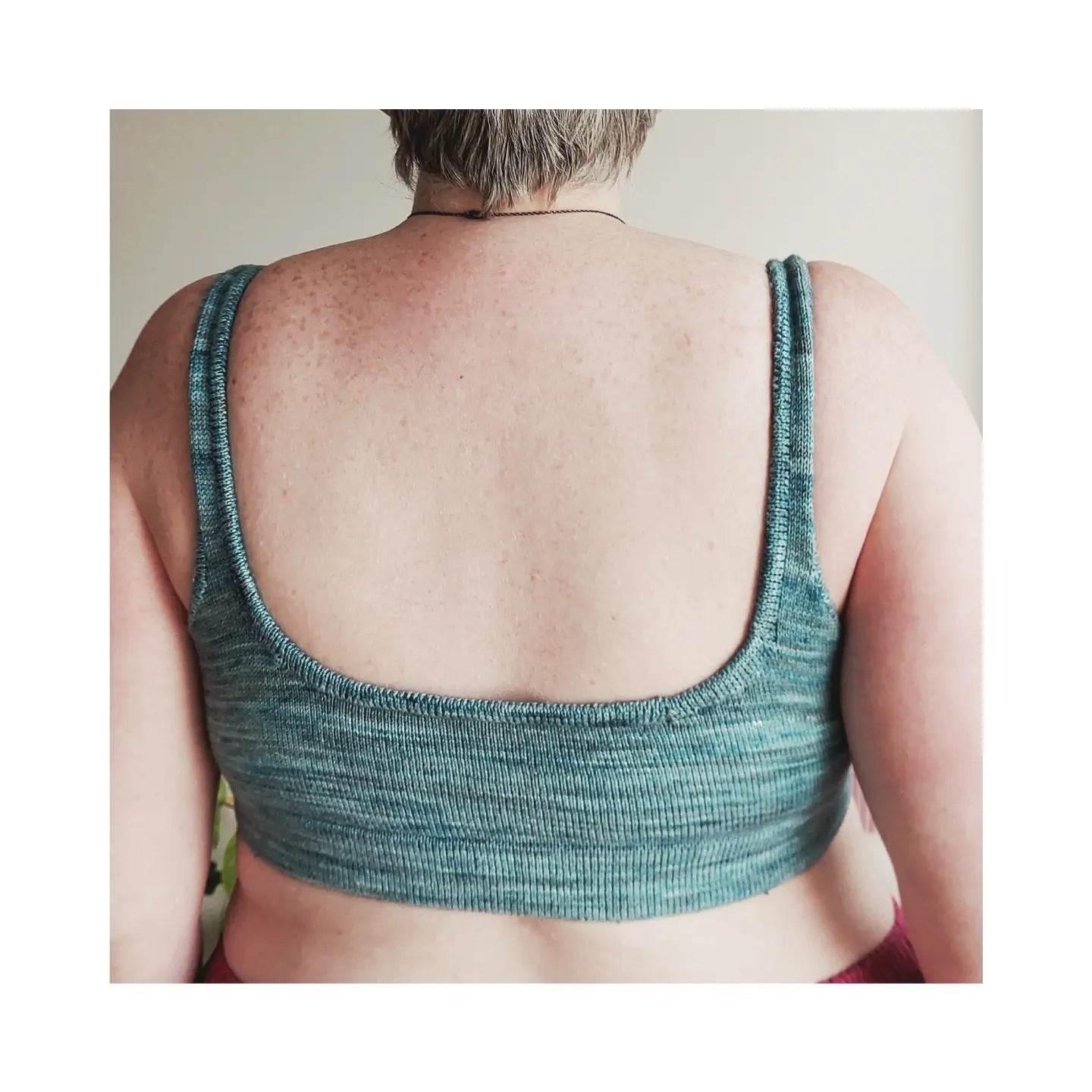 Seen from behind, Jenny wears a hand knit bralette, made with grey-green variegated yarn.