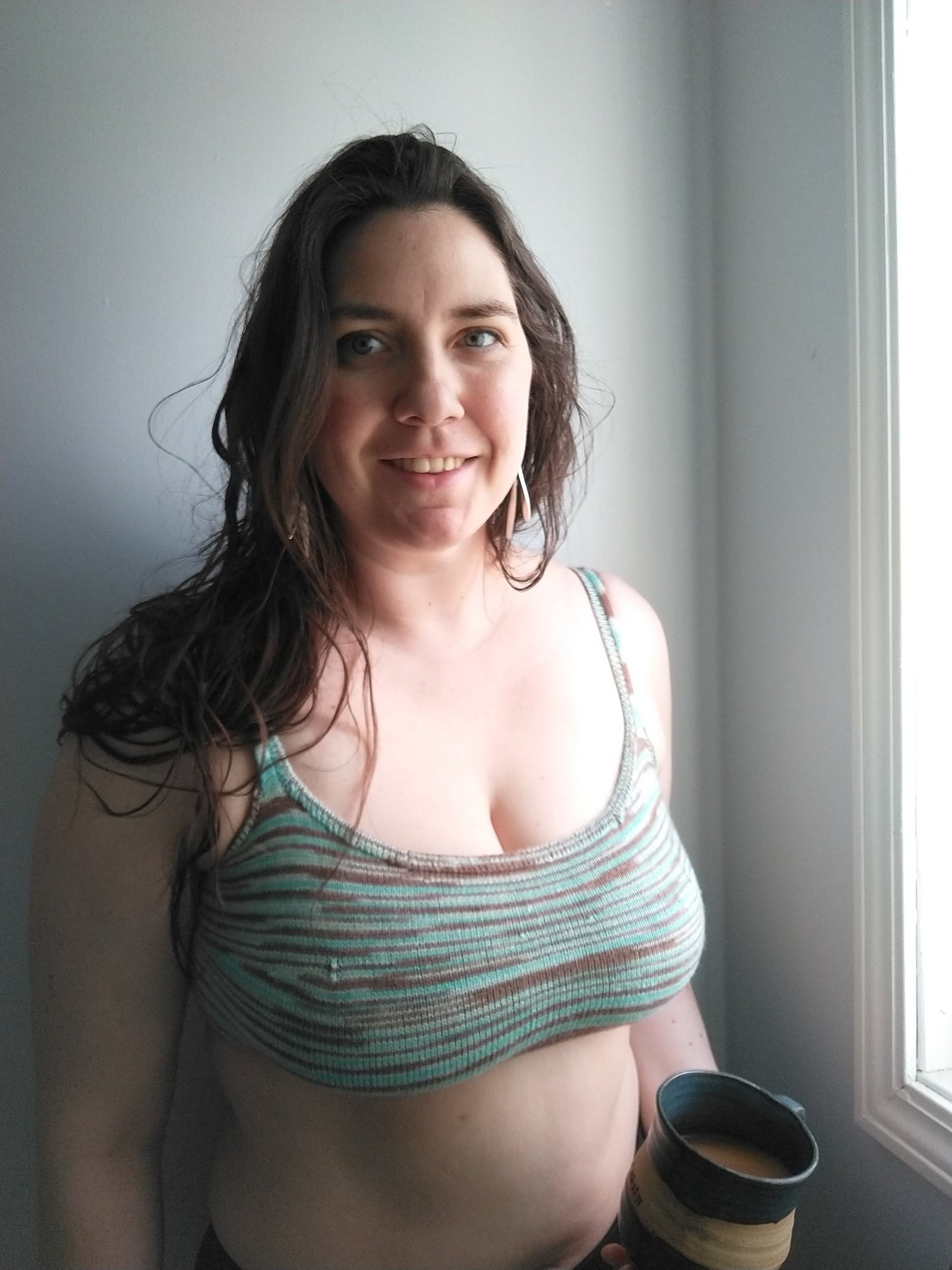 Gillian holds a mug of tea, smiling at the camera. She wears a tailored bralette, knit with teal and brown variegated yarn.
