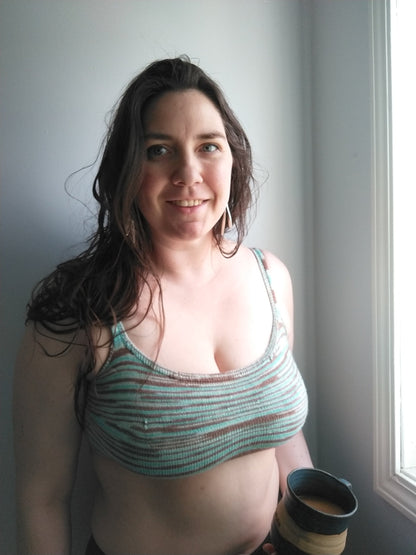 Gillian holds a mug of tea, smiling at the camera. She wears a tailored bralette, knit with teal and brown variegated yarn.