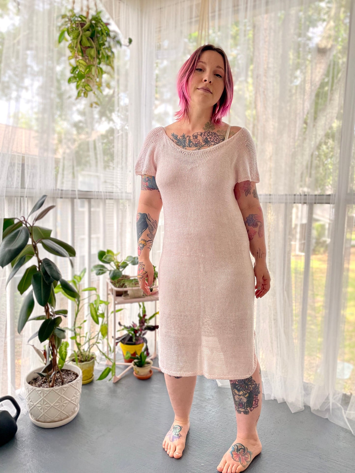 Bess stands in a room, smiling at the camera. She wears a white, hand knit dress with short sleeves.