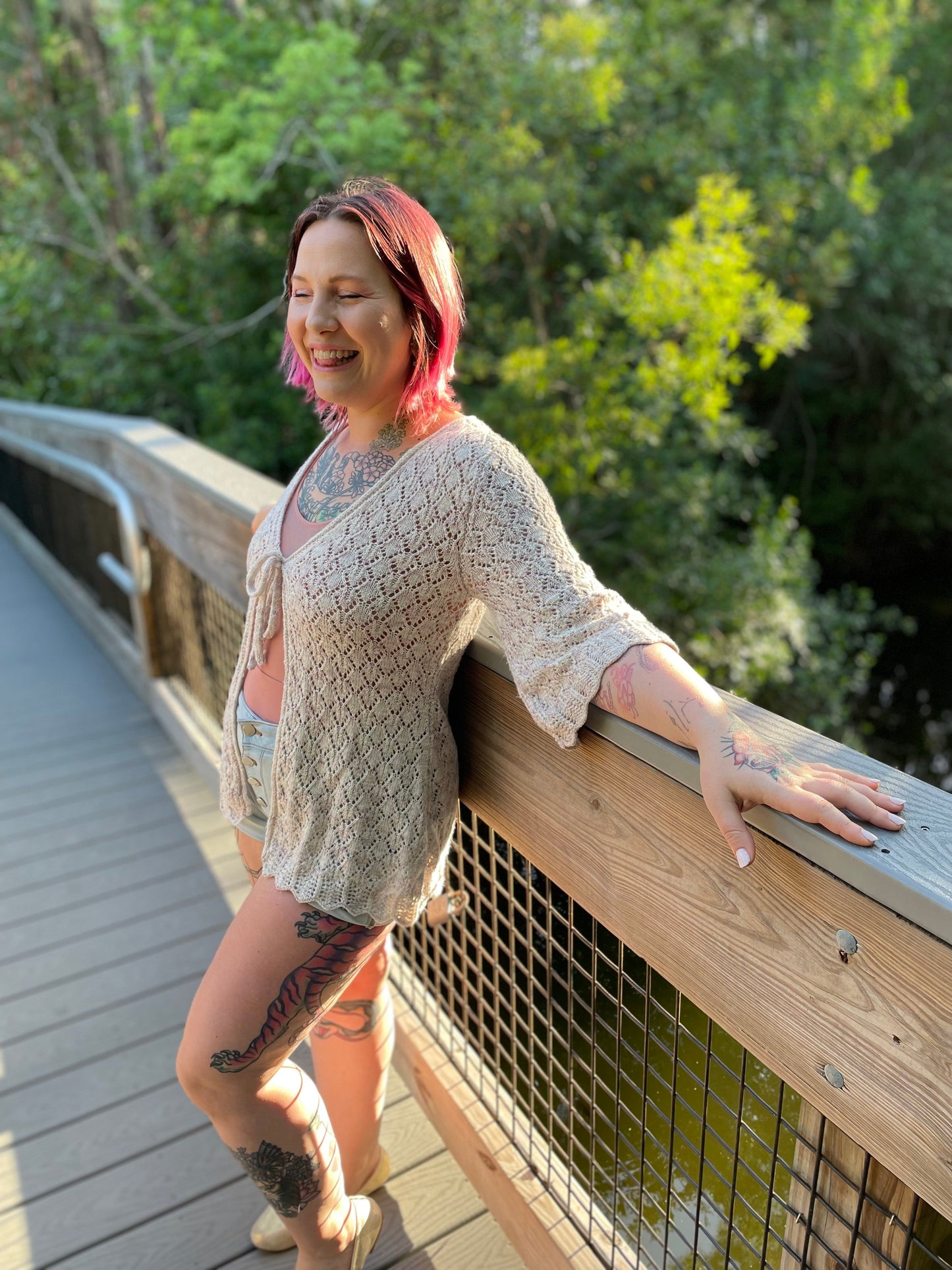 Seen from the side at a 3/4 angle, Bess leans against a wooden bridge railing. She wears a white cardigan, knit in a lace stitch with 3/4 length flared sleeves and a scalloped hem. The front is closed with a simple tie.