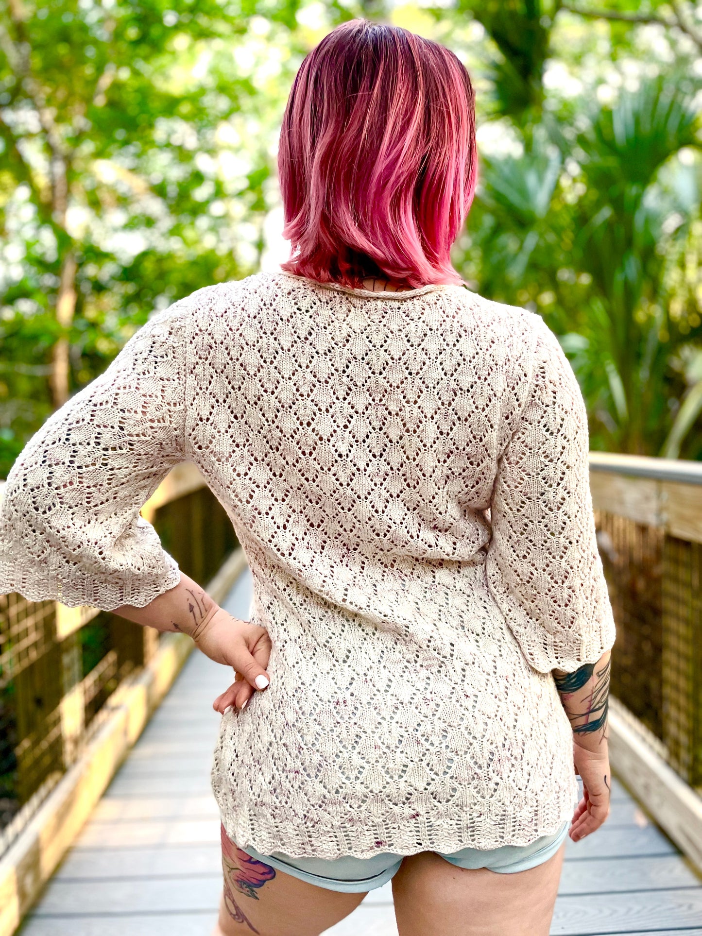 Seen from behind, Bess wears a white, light weight cardigan over denim shorts. The lacy cardigan features flared, 3/4 length sleeves and a scalloped hem. 