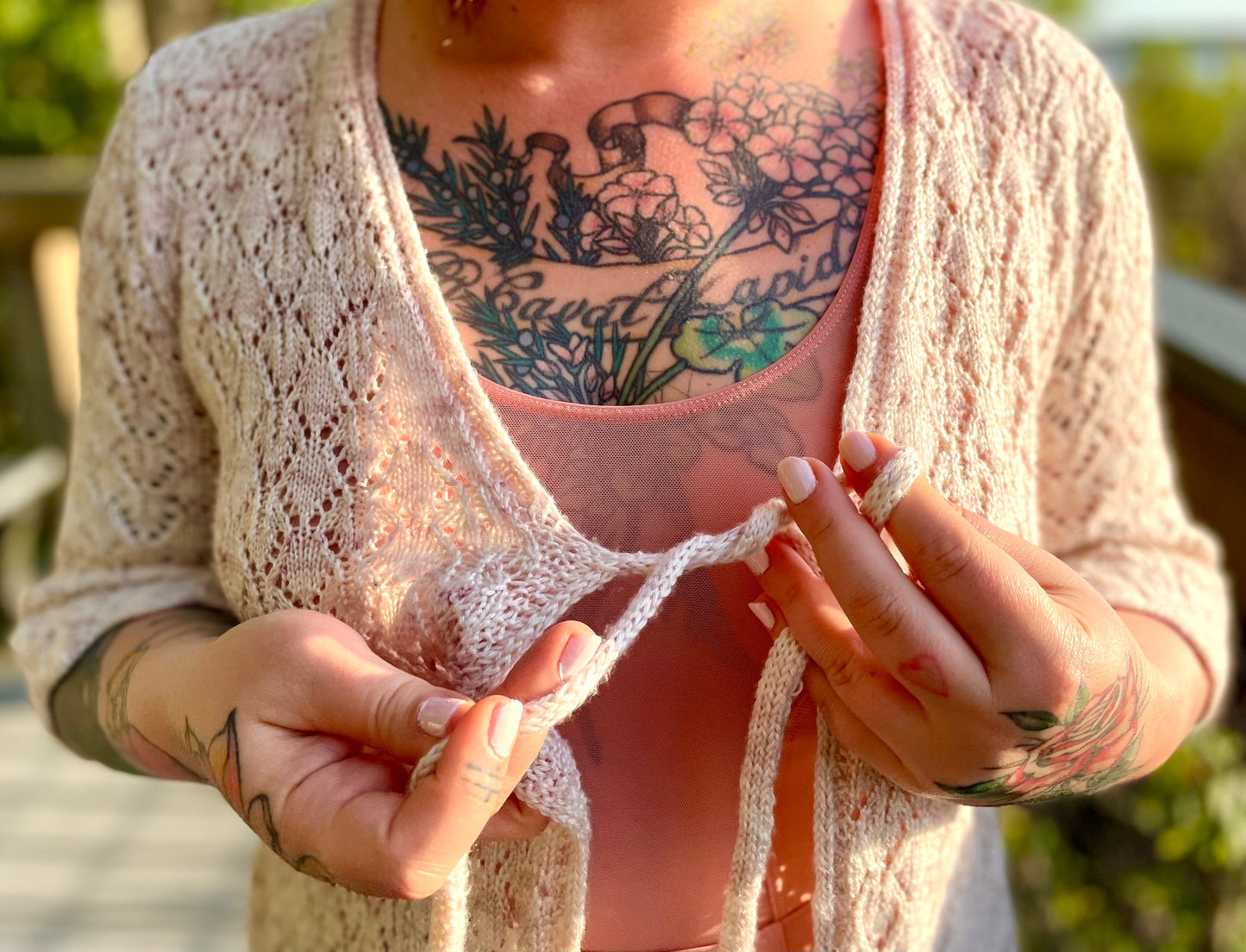 Seen close up, Bess wears a white cardigan knit in a delicate lace stitch. She ties the front strings over a light pink tank.