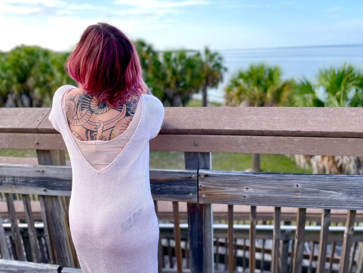 Bess leans against a bridge railing, her back to the camera. She wears a knit white dress, the back scooped to show off her tattoos.