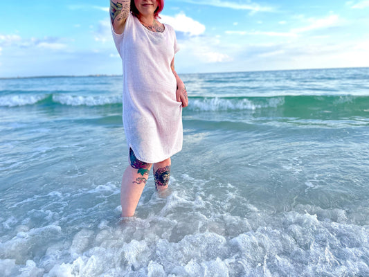 Bess stands on the shore, waves coming up around her shins. She wears a hand knit dress with short sleeves and a scoop neck, made with white yarn.