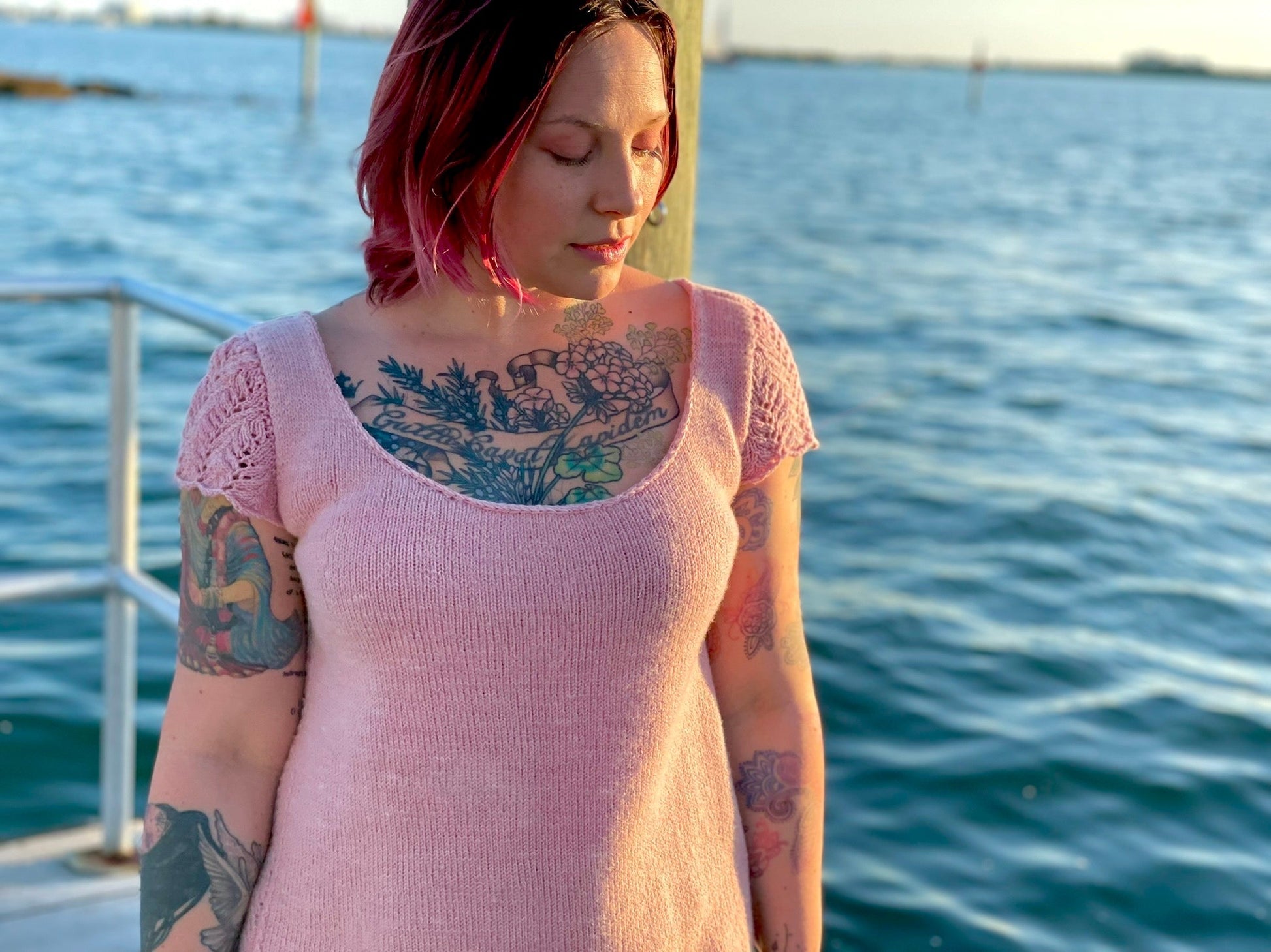 A woman stands on a dock wearing a hand knit tee in baby pink fingering weight organic wool, it has lace sleeve caps and a generous scoop neckline.