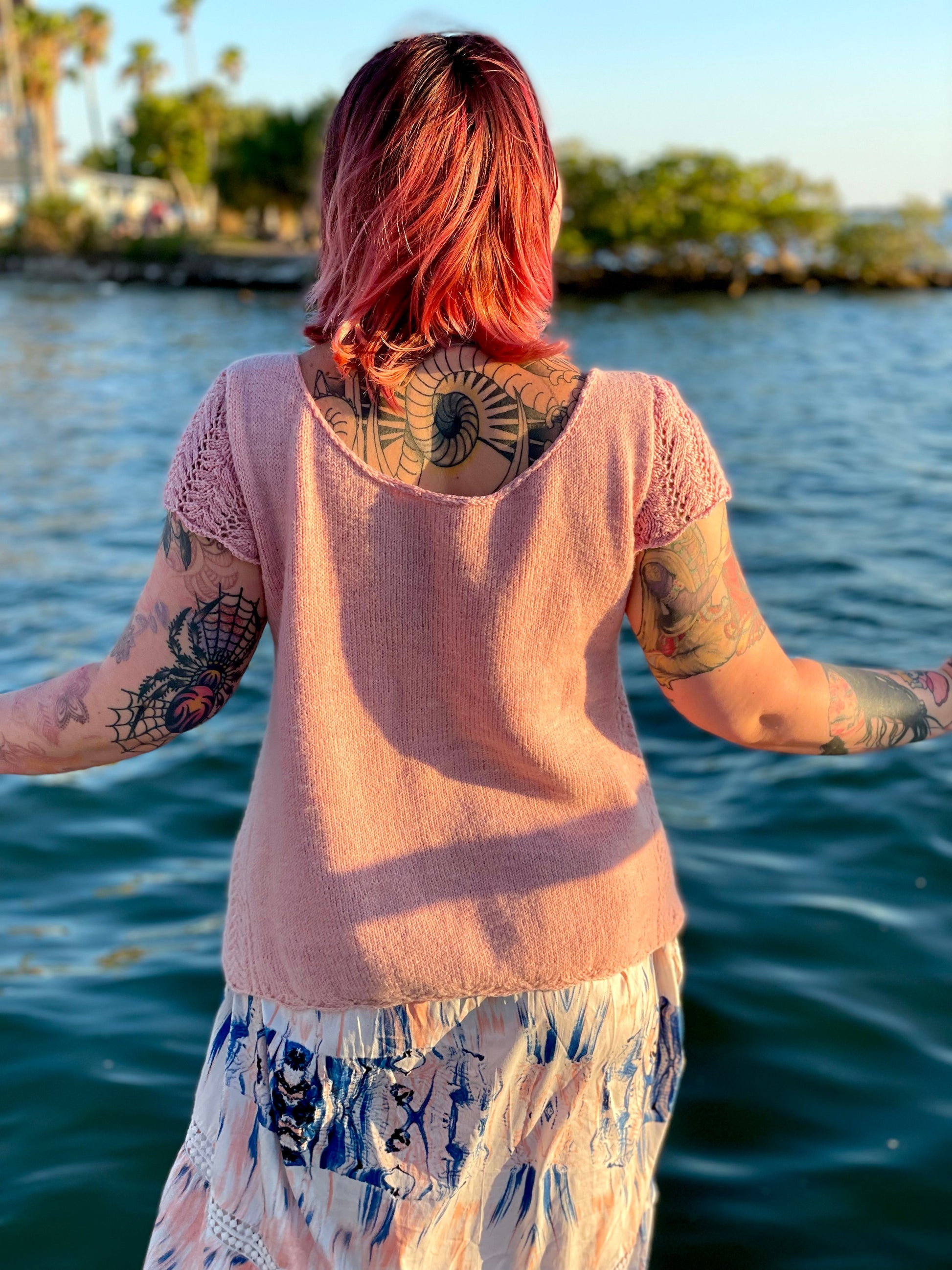 A white woman with rose gold hair and many visible tattoos stands on a docking facing away from the camera. She looks out over the water wearing a hand knit wool tee. The anna tee knitting pattern features lace cap sleeves, generous positive ease and a scooping front and back neckline.