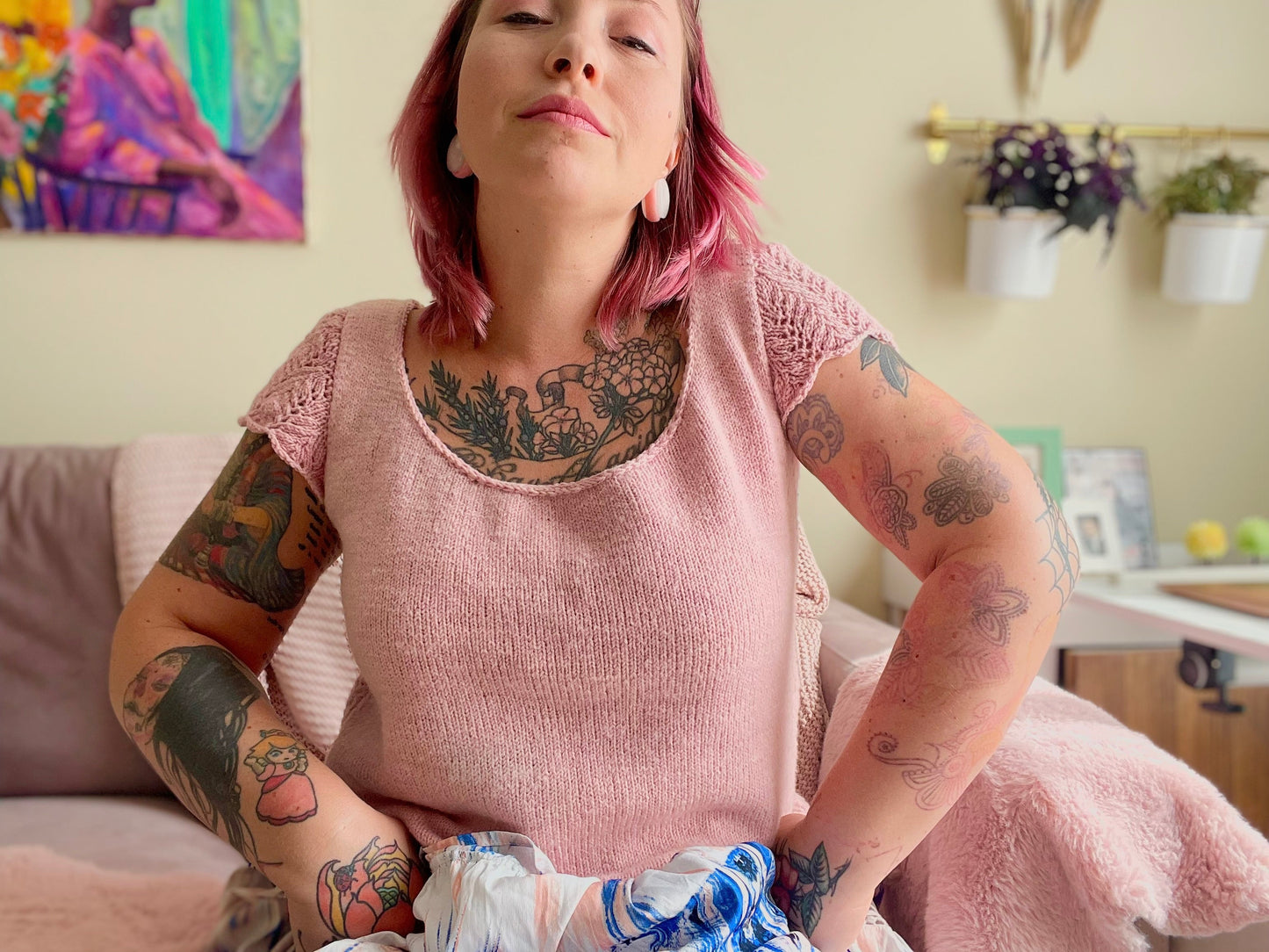 A woman sits on a dusty pink couch wearing a light pink wool tee with lace cap sleeves. She has many visible tattoos showing and sits with her hands on her hips and her shoulders shrugged in a defiant gesture.