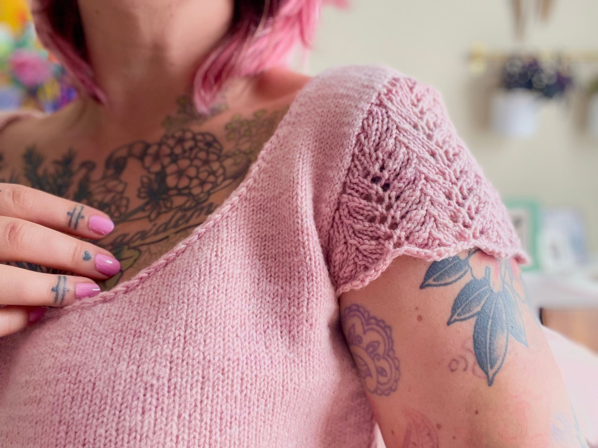 A close up shot of a baby pink hand knit wool tee with lace sleeve caps and a striking plunging neckline. A white woman is wearing this sweater tee, she has many visible tattoos showing on her chest, arms and hands.