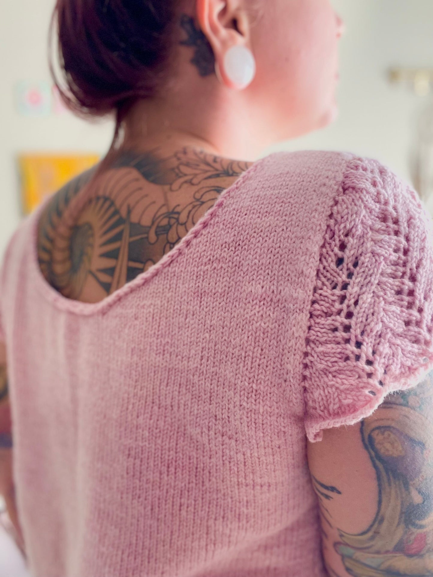 A close up shot of a hand knit pink t-shirt with lace cap sleeves and a scooping back neckline. The white woman wearing the tee has her pink hair in a bun revealing a sprawling back tattoo.