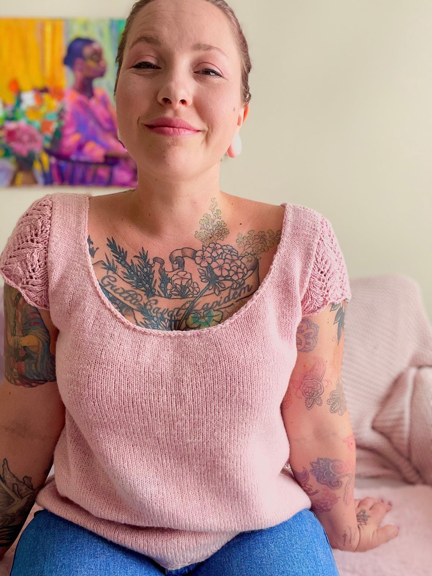 A white woman with many visible tattoos sits on a dusty pink couch. She wears a hand knit wool tee in hand dyed baby pink fingering weight yarn with lace sleeve caps and a striking scoop neckline.