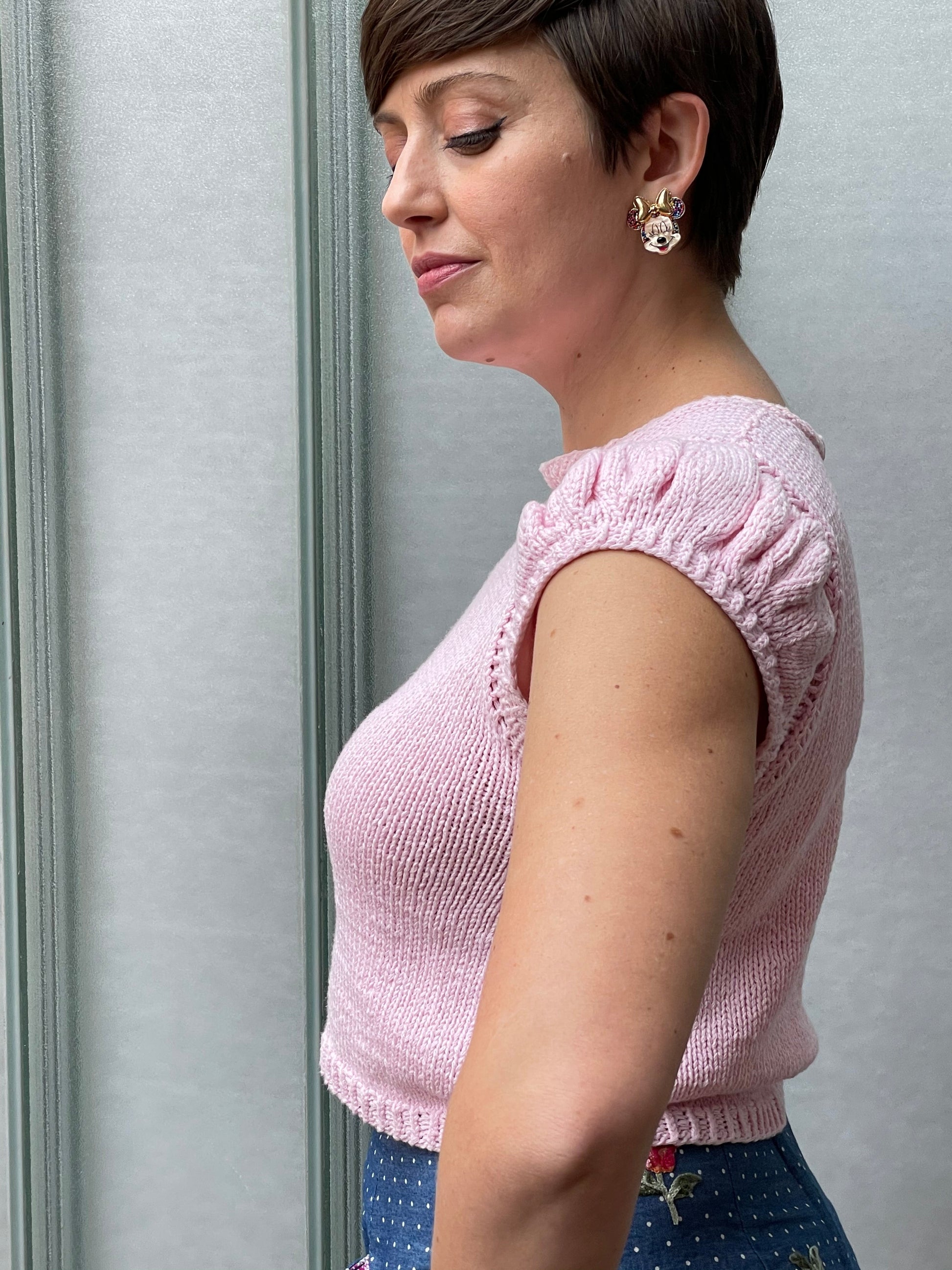Seen from the side, Jeanette wears a light pink tee, knit with ruffled sleeves, over a blue skirt. The tee is a cropped, waist length.