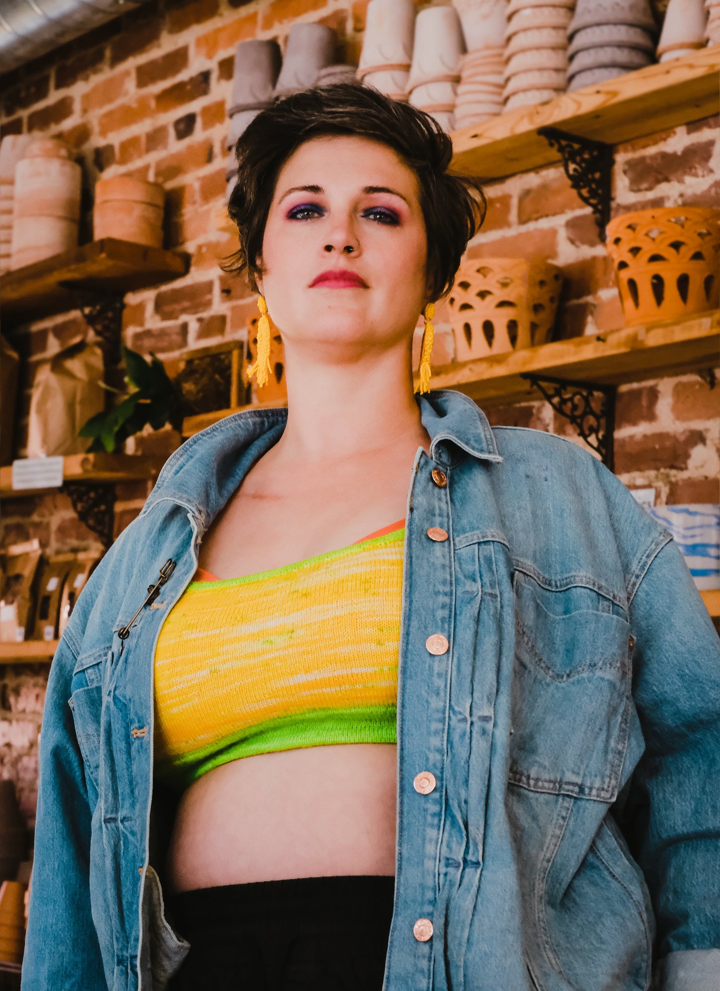 Jen looks down at the camera, wearing a denim jacket and black leggings with a hand knit bralette. The bralette is made with bright yellow yarn and is knit with a neon green band.