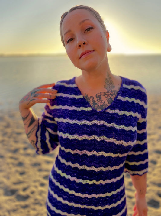 Bess stands on the shoreline, looking at the camera. She wears a blue and white striped dress, knit in a feather and fan stitch with 3/4 length sleeves.