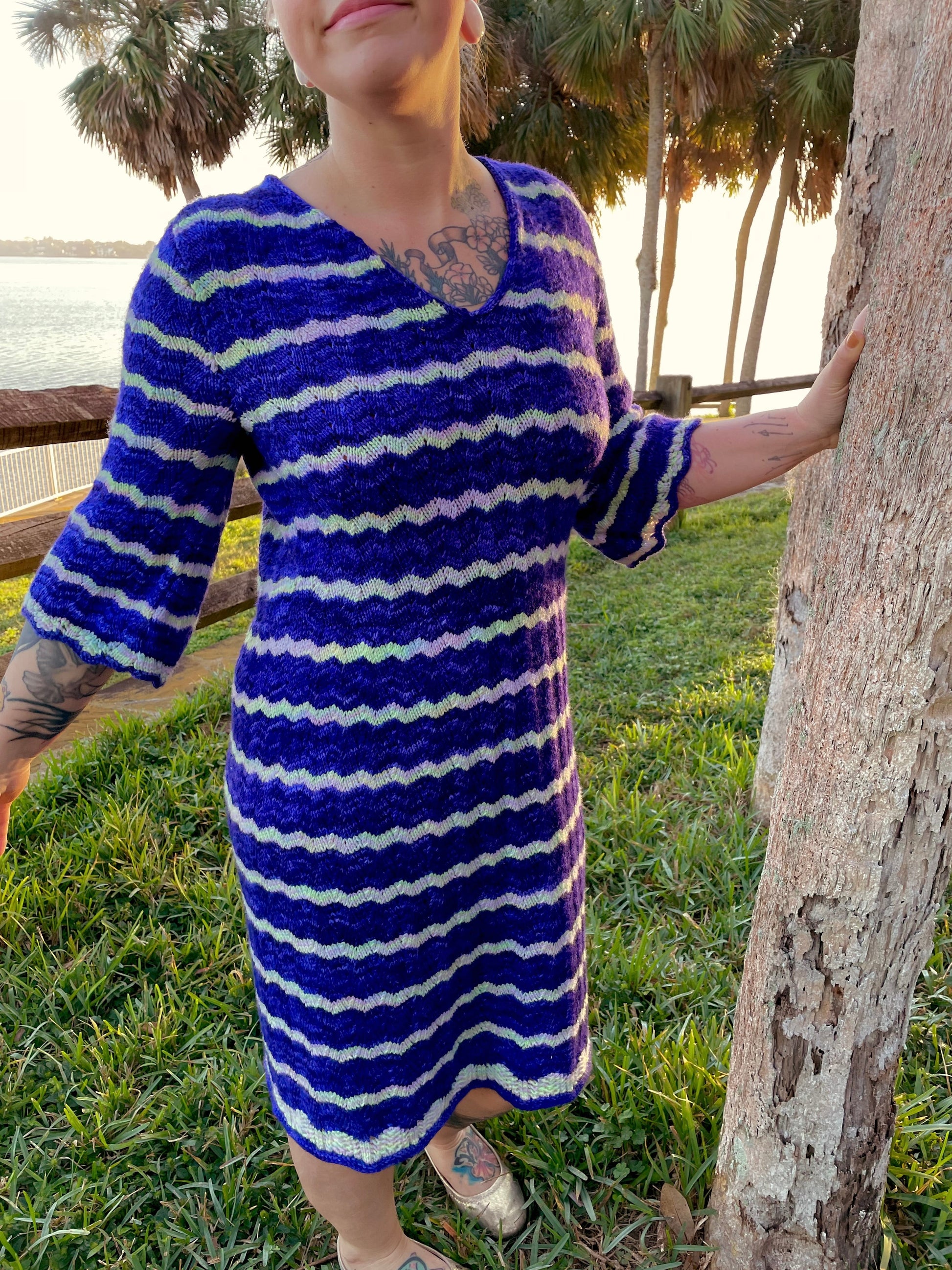 Seen from the neck down, Bess wears a lace knit dress, made with blue and white stripes and a gentle V-neckline. Palm trees can be seen in the background.