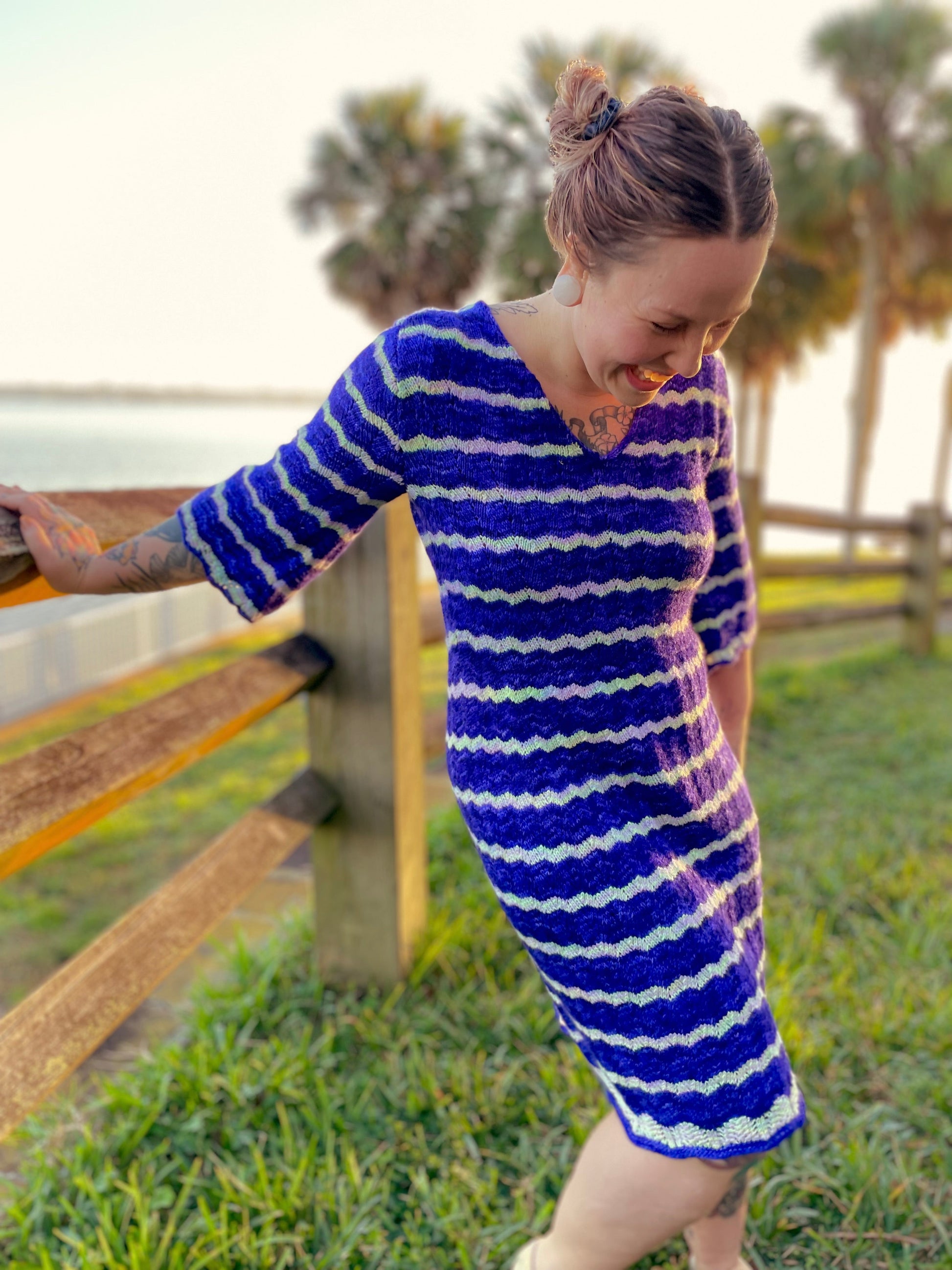 Bess stands, laughing, next to a wooden fence on the shoreline. She wears a hand knit blue and white dress that falls to her knees.