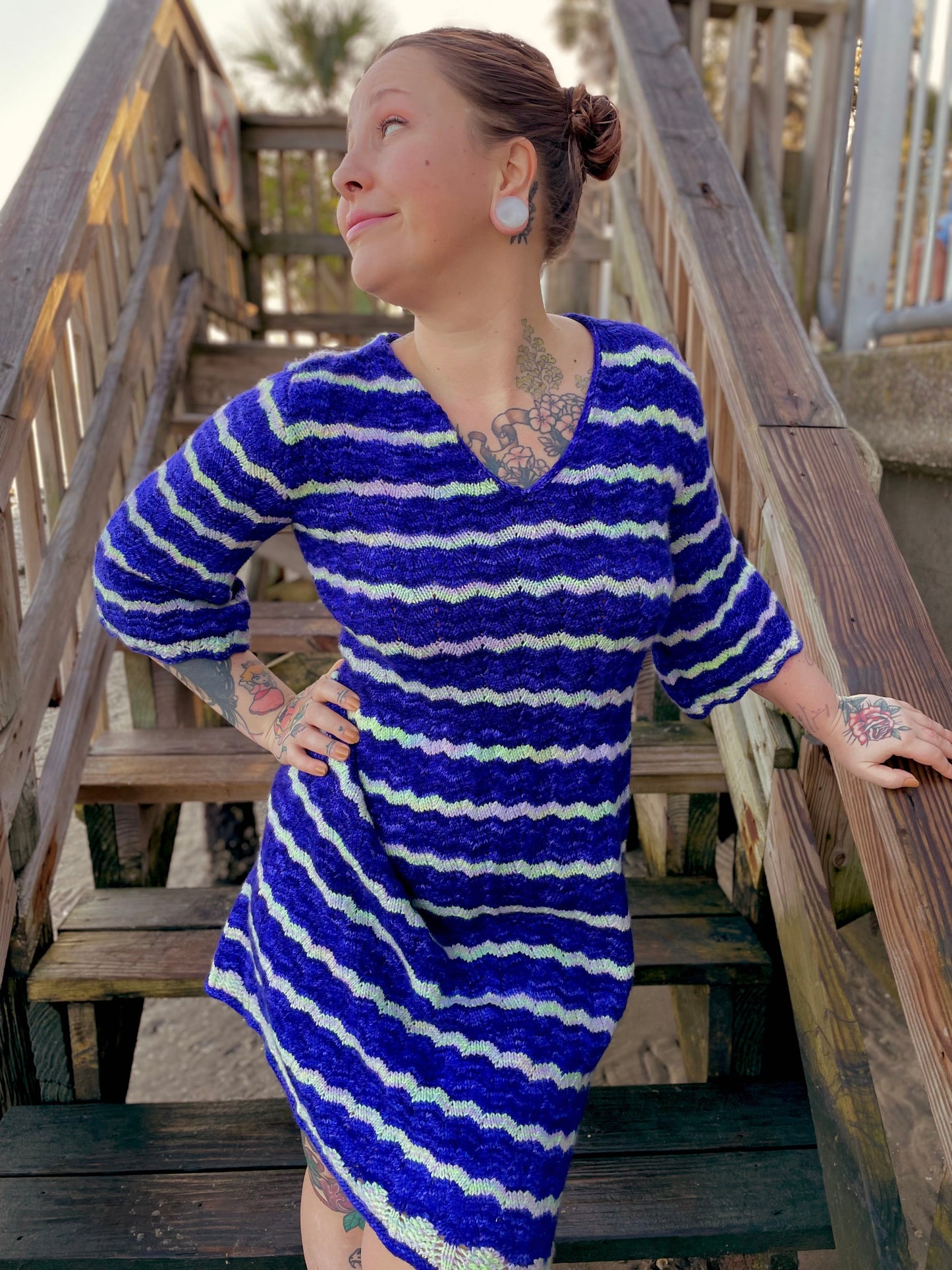 Bess stands on a deck staircase, looking off camera with one hand on her hip. She wears a blue and white dress, knit with a feather and fan stitch and 3/4 length sleeves.