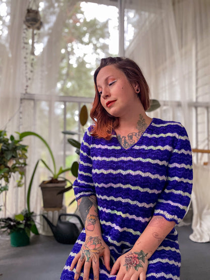 Bess sits inside, looking to the side. Her blue and white lace dress is knit with a gentle V-neckline and 3/4 length sleeves.