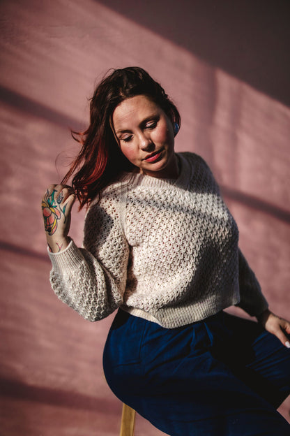 Bess sits on a stool, wearing blue pants with a cream coloured lace knit sweater, the Mary Raglan. She is looking down towards the bottom of the frame.