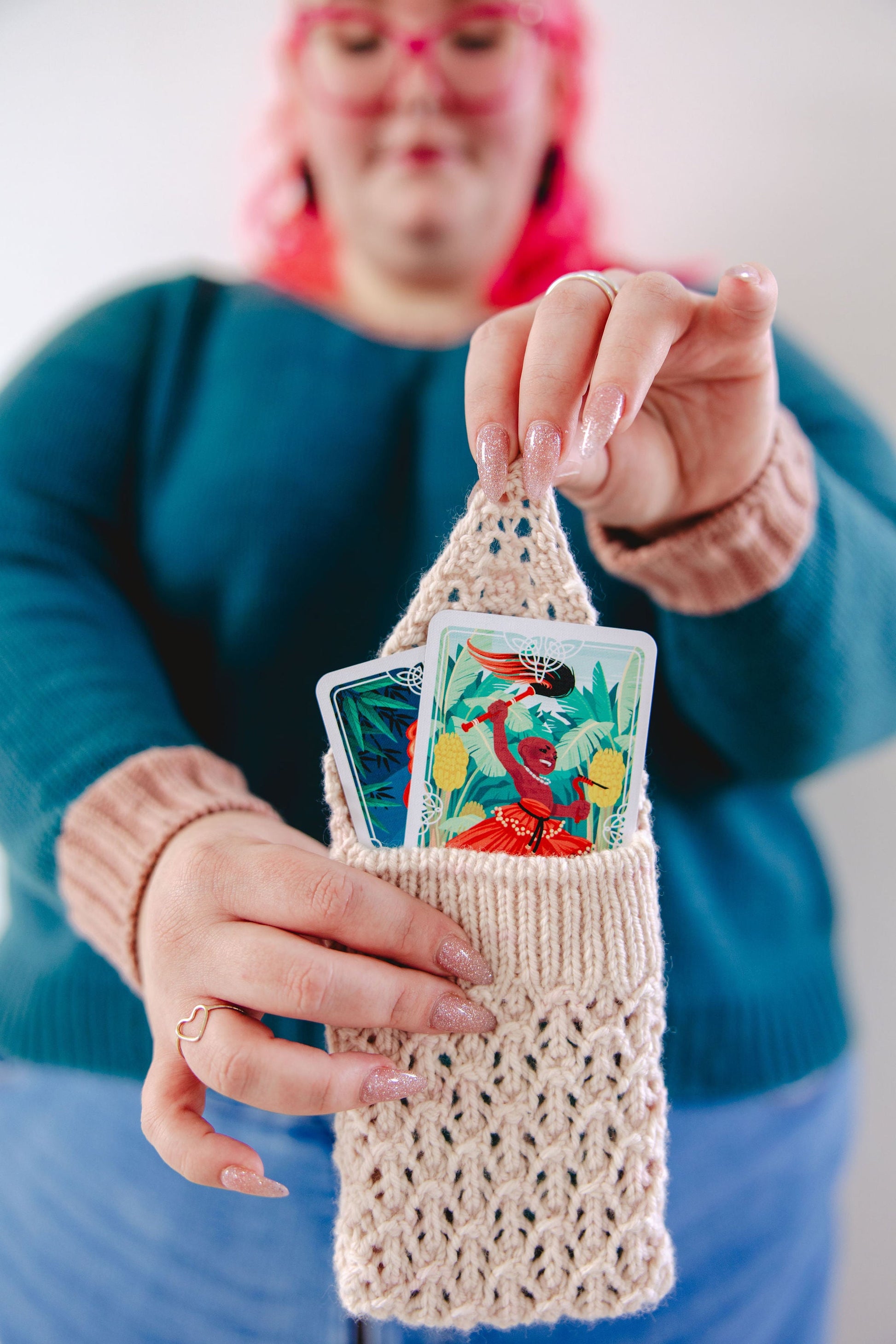 Courtney, who has pink hair and matching pink glasses, holds a lace knit pouch open. Inside the pouch are two brightly coloured tarot cards.