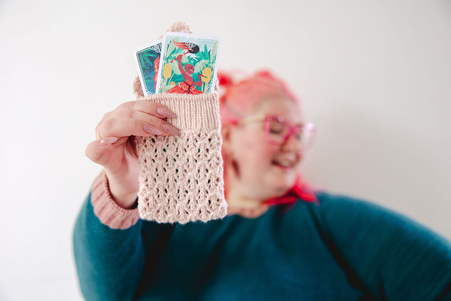 Courtney, who has pink hair, holds a cream colored lace knit pouch, the Mary Tarot Pouch. The pouch holds two brightly colored tarot cards.