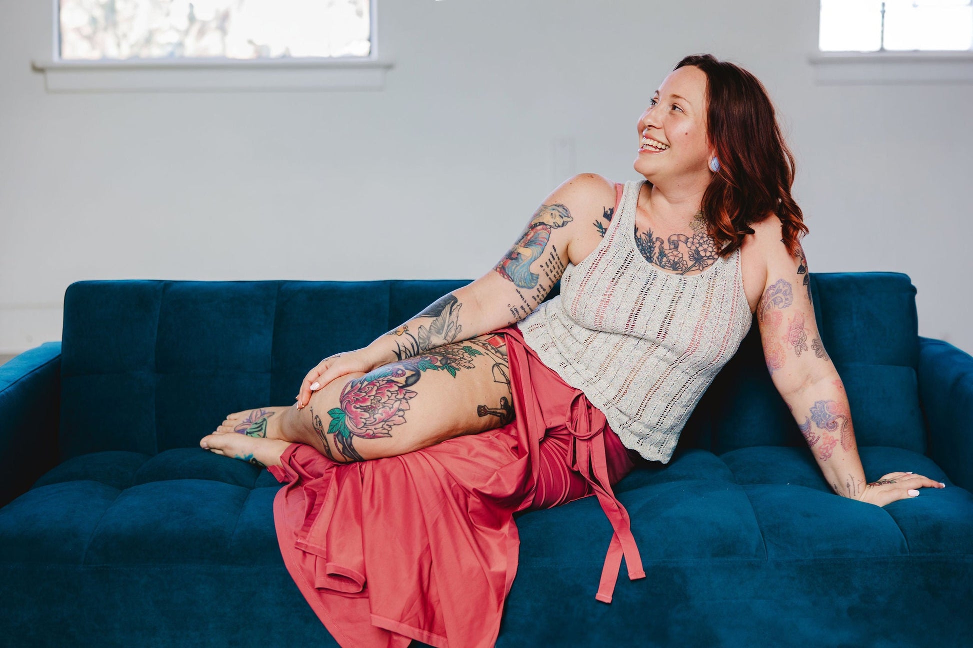 Smiling and looking off camera, Bess reclines on a couch. She wears a chevron lace knit tank, the Tori Tank, with a flowy pink skirt.