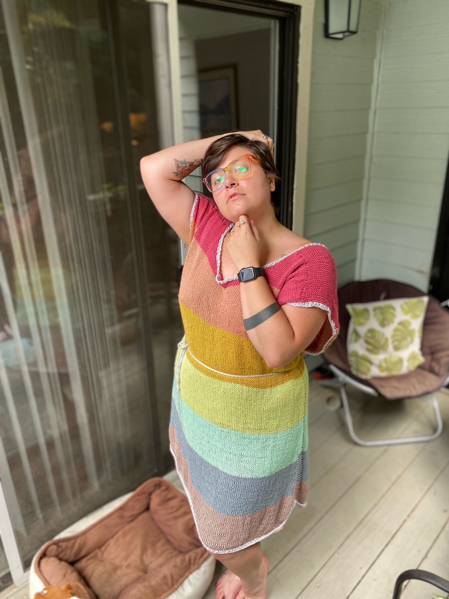 A person stands on their porch, smiling at the camera. Her hand knit dress is knit in rainbow sections.