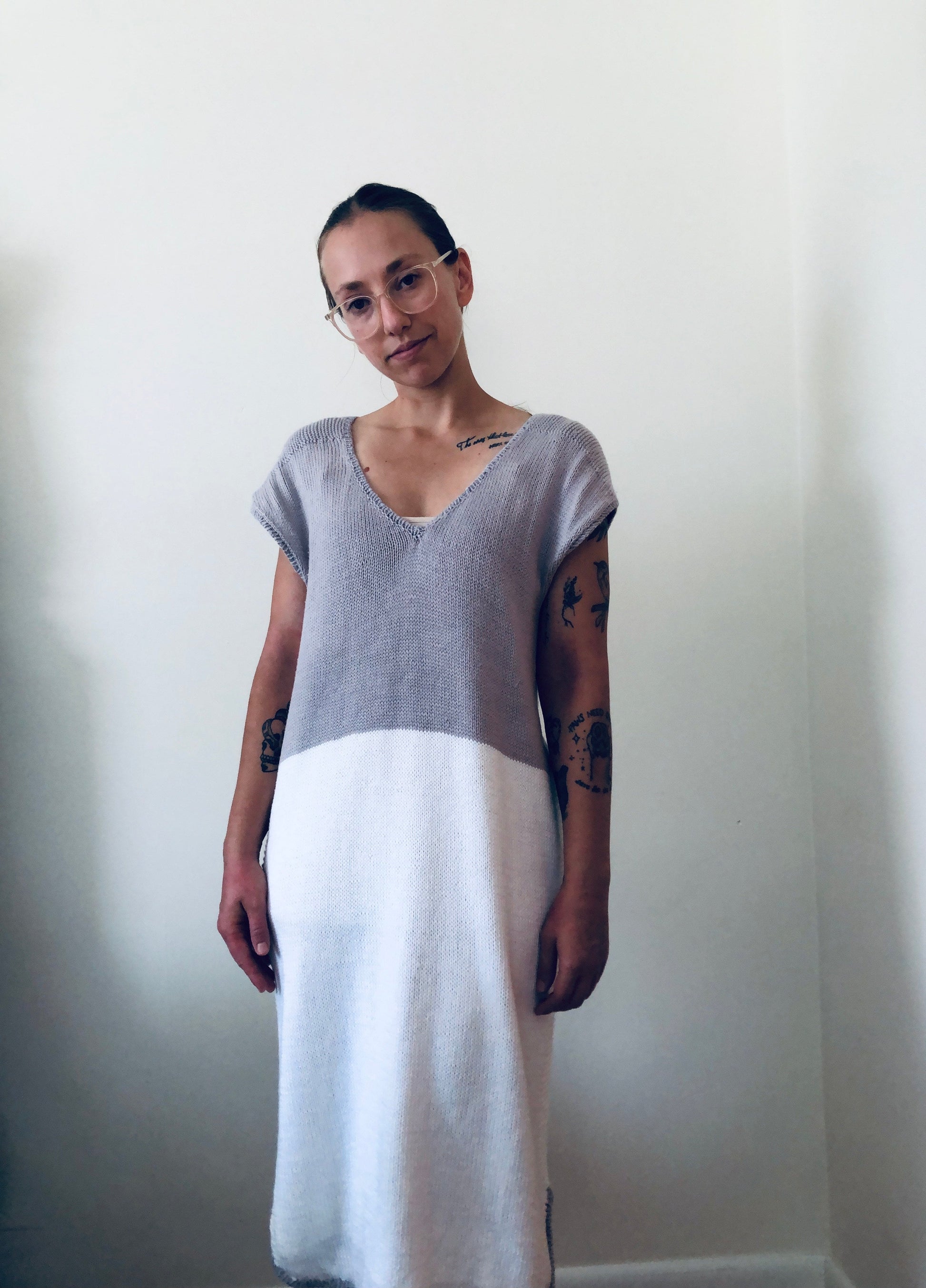 A woman stands, smiling at the camera, wearing a white and grey hand knit dress. The dress has a scoop neck and short sleeves.