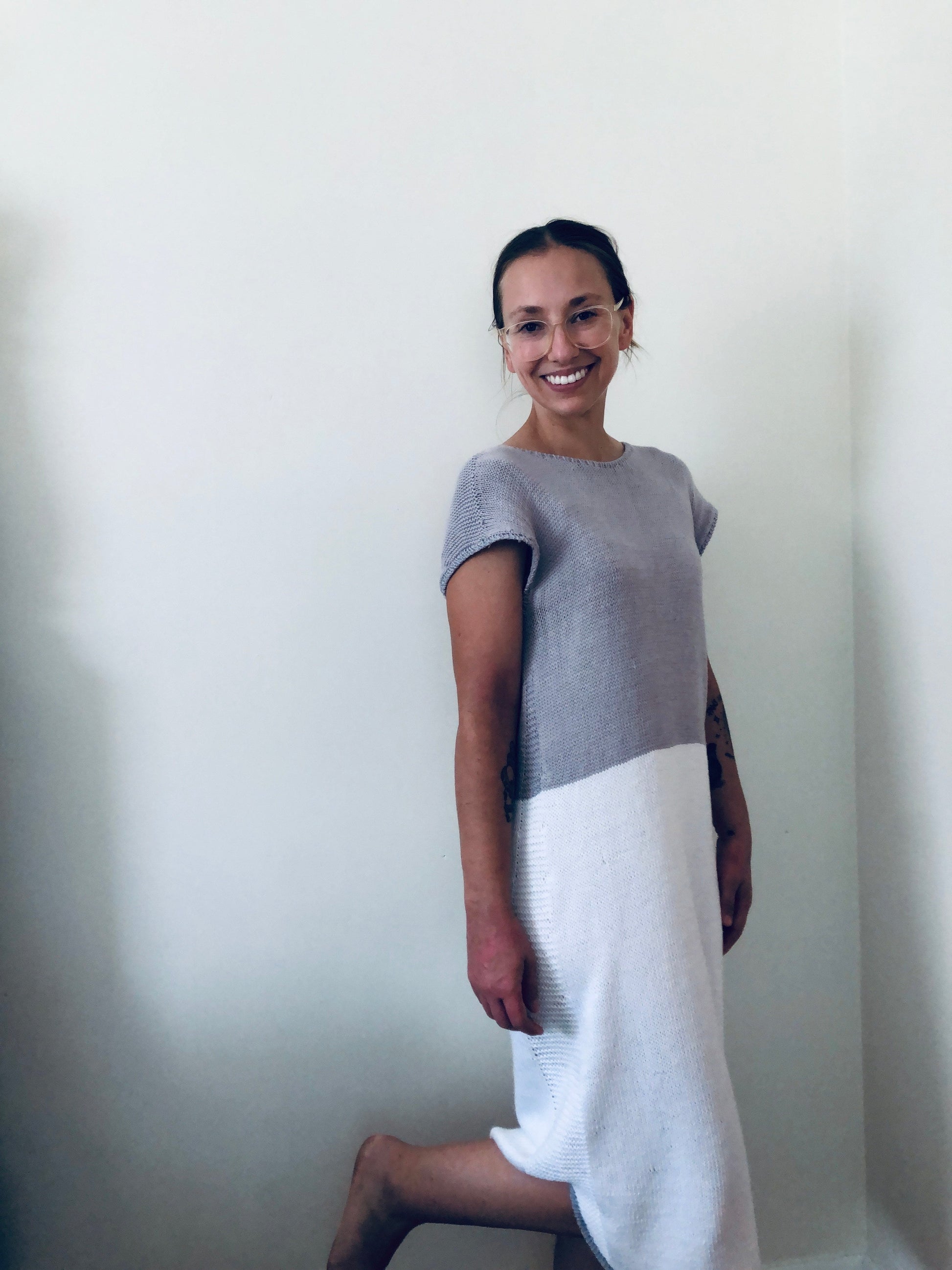 Seen from a three quarter angle, a woman smiles at the camera. She wears a grey and white hand knit dress.